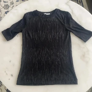 Svart plisserad topp från H&M - Säljer en svart plisserad topp från H&M med rund halsringning och korta ärmar. Toppen har en snygg struktur i tyget som ger en modern känsla. Perfekt att styla till olika outfits!