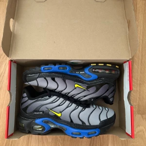 Nike Air Max Plus med blå och gula detaljer - Säljer ett par Nike Air Max Plus med svart och grå ovandel, blå sula och gula detaljer. Skorna har snörning, rund tå och synliga Air Max-enheter i sulan. Perfekta för dig som vill sticka ut med en sportig och modern look.