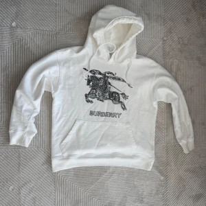 Vit hoodie från Burberry med tryck - Säljer en vit hoodie från Burberry med ett stort svart tryck av en riddare på häst och texten 'BURBERRY' framtill. Tröjan har huva med dragsko och en stor magficka. Perfekt för dig som gillar stilrena märkesplagg.