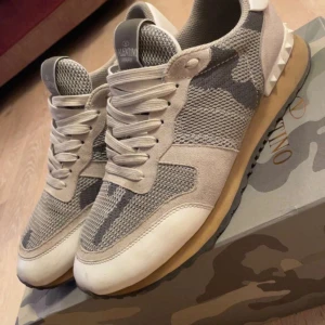 Valentino Rockrunner sneakers i beige och grå - Snygga Valentino Rockrunner sneakers i en cool beige och grå camouflage-design. Skorna har snörning och är tillverkade i en kombination av mocka och syntetmaterial. Perfekta för en stilren och avslappnad look.