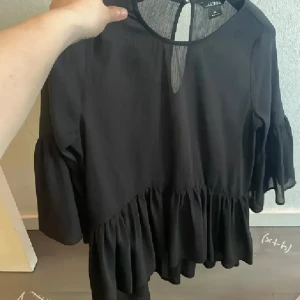 Svart volangblus  - Säljer en svart blus från Jacquelin de Yong med volang nertill och trekvartsärmar. Blusen har rund halsringning och en liten öppning bak i nacken. Tunt och luftigt material som ger ett snyggt fall.