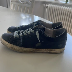 Svarta golden goose - Säljer ett par svarta sneakers med vita snören och vit sula. Skorna har en klassisk design med rund tå och platt sula. Perfekta för dig som gillar en enkel och stilren look till vardags.