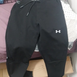 Svarta mjukisbyxor från Under Armour - Svarta mjukisbyxor från Under Armour med elastisk midja och dragsko. Byxorna har logga på benet och bred resår med texten 'UNDER ARMOUR' runt midjan. Perfekta för träning eller chill.