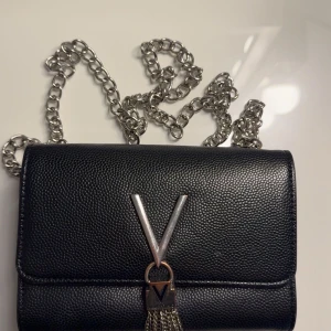 Svart axelväska från Valentino med kedja - Säljer en ÄKTA svart axelväska från Valentino med silverfärgad kedja och dekorativt V-spänne framtill. Väskan har en elegant tofsdetalj i metall och är tillverkad i ett strukturerat syntetmaterial. Som ni kan se är låset rostigt men jag tror man kan knipsa upp det om man vill.💕