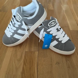 Grå Adidas Campus sneakers - Säljer ett par grå Adidas Campus sneakers med vita ränder och vit sula. Skorna har klassisk Adidas-logga på plösen och är tillverkade i mocka med vita snören. Perfekta för dig som gillar stilrena och tidlösa sneakers.