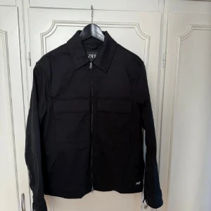Svart overshirt från ZARA - Säljer en svart overshirt från ZARA med dragkedja framtill och två bröstfickor. Jackan har klassisk krage och lång ärm, perfekt för lager-på-lager-stil. Enkel och stilren design som passar till många outfits. Jackan är som ny. Pris kan diskuteras vid snabb affär.