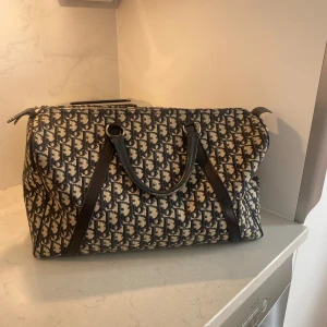 Mönstrad axelväska från Dior - Säljer en klassisk axelväska från Dior med blått och beige monogrammönster. Väskan har svarta skinnhandtag och detaljer samt dragkedja upptill