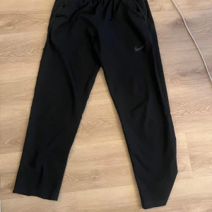 Nike DRI FIT byxor - Svarta mjukisbyxor från Nike med diskret logga på vänster lår. Byxorna har resår i midjan och raka ben, perfekta för en avslappnad stil eller träning. Mjukt material och bekväm passform.