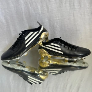 Adidas F50 Adizero fotbollsskor svart/guld - Snygga Adidas F50 Adizero fotbollsskor i svart med vita ränder och guldiga detaljer. Skorna har snörning, platt sula med dobbar och är tillverkade i syntetmaterial. Guldiga detaljer på hälen och sulan ger en riktigt exklusiv look. Perfekta för dig som vill sticka ut på planen.