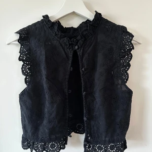 Svart broderad väst med spetsdetaljer - Supersnygg svart väst med broderade blommor och spetskant vid ärmhål och nederkant. Västen har små knappar framtill och en söt volangkrage. Från COS i storlek XS. 