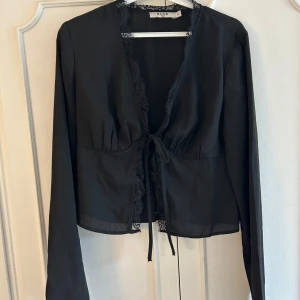 Svart blus med spets från NA-KD - Säljer en svart blus från NA-KD med långa ärmar och v-ringning. Blusen har fina spetsdetaljer längs kanten och knyt framtill. Tunn och flowy känsla, perfekt för att lyfta din outfit med en elegant touch.