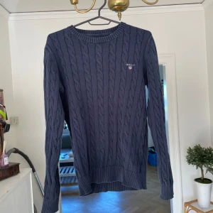Tröja Gant - Kabelstickad mörkblå tröja från GANT i premium bomull. Klassisk rund halsringning, ribbade muddar och diskret logga på bröstet. Perfekt för dig som gillar preppy stil och vill ha en tidlös basic i garderoben. Tyvärr en fraktur i höger armbåge men märks ej.