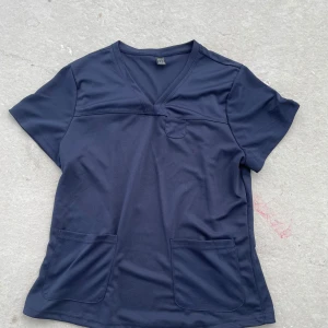 Mörkblå v-ringad kortärmad topp - Mörkblå kortärmad topp med v-ringning och tre praktiska fickor framtill. Tillverkad i mjukt och lätt polyestertyg som känns skönt mot huden. Enkel och clean design som passar perfekt till jeans eller leggings.