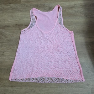 Ljusrosa ärmlös linnetopp med spets - Supersöt ljusrosa linnetopp med broderad spets framtill och slät baksida. Toppen är ärmlös och har en lätt, luftig känsla – perfekt för varma dagar. Spetsen har ett fint bladmönster som ger en romantisk vibe. Passar dig som gillar en feminin och chill stil.