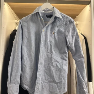 Ljusblå skjorta från Polo Ralph Lauren - Säljer en klassisk ljusblå skjorta från Polo Ralph Lauren i custom fit. Skjortan har lång ärm, knappar framtill och den ikoniska broderade loggan på bröstet. Perfekt för dig som gillar stilrena och tidlösa plagg. Finns en liten reva i tyget som syns på bilderna 