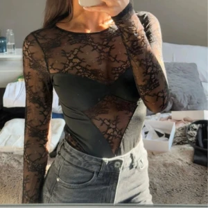 Svart spetsig långärmad body - Säljer en svart långärmad body med transparent spets och coola detaljer. Toppen har en figurnära passform och är perfekt för dig som vill sticka ut. Snygg att styla med jeans eller kjol för en trendig look.