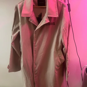 Undercover Witch embroidery coat SS17 - Säljer en beige trenchcoat från Undercover med cool pixelgrafik av en häxa på ryggen och broderad logga på bröstet. Jackan har klassisk krage, knäppning framtill och fickor i sidorna. Perfekt statement-piece för dig som gillar unika plagg.