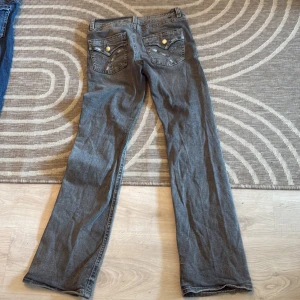 Gråa bootcut jeans från Gina Tricot - Säljer ett par gråa jeans från Gina Tricot med bootcut-modell. Jeansen har snygga detaljer på bakfickorna med knappar och klassisk femficksdesign. Tillverkade i mjukt jeanstyg och passar perfekt till en avslappnad stil.