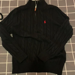 Svart cardigan stickad tröja Polo Ralph Lauren - Svart kabelstickad tröja från Polo Ralph Lauren med halv dragkedja och hög krage. Klassisk röd logga broderad på bröstet. Tröjan är långärmad och har ribbade muddar och nederkant. Perfekt för lager-på-lager under kyliga dagar.