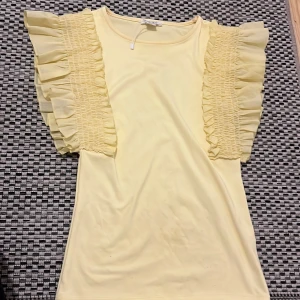 Gul volangtopp från Amisu - Supersöt ljusgul topp från Amisu med breda volangärmar och smockdetaljer längs axlarna. Toppen har rund halsringning och är tillverkad i ett lätt, mjukt material som känns skönt mot huden. Perfekt för dig som vill sticka ut med en feminin och trendig look.