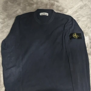 Mörkblå Stone Island tröja i bomull - Snygg mörkblå långärmad tröja från Stone Island med klassisk patch på vänster ärm. Tröjan är tillverkad i mjuk bomull och har en enkel, stilren design med rund hals. Perfekt för dig som gillar streetwear och vill ha något stilrent i garderoben.