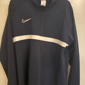 Marinblå Nike half zip tröja - Snygg marinblå tröja från Nike med vit rand över bröstet och klassisk Nike-logga. Tröjan har half zip och långa ärmar, perfekt för dig som gillar sportig stil. Skön passform och enkel att matcha med jeans eller träningsbyxor.