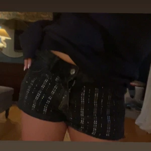 Svarta glittriga jeansshorts - Säljer ett par svarta jeansshorts med glittriga detaljer på. Shortsen är låga i midjan och har klassisk femficksdesign med knapp och dragkedja framtill. Köpta i Milano!