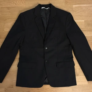 Svart kavaj från H&M, stl 158 - Klassisk svart kavaj från H&M i storlek 158. Kavajen har två knappar framtill, två fickor med lock och en bröstficka. Snyggt figursydd modell med slät finish och helfodrad insida. Perfekt för dig som vill ha en stilren look.
