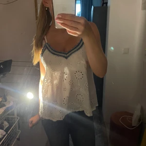 Vit  linnetopp med spets - Supersöt vit linnetopp med broderade blommor och hålmönster. Toppen har v-ringning med blå och vit spetskant och tunna axelband. Den är luftig och perfekt för varma dagar, 