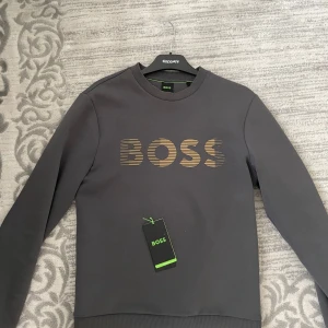 Mörkgrå BOSS sweatshirt stretch  - Snygg mörkgrå sweatshirt från BOSS i storlek XS. Tröjan har ett stilrent guldigt BOSS-tryck på bröstet och klassisk rund hals. Perfekt basic med lång ärm och mjukt material som känns skönt mot huden. Ny pris 1599kr se ovan, frakt /swish eller mötas 