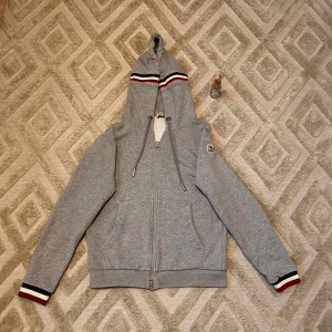 Grå hoodiejacka från Moncler - Grå hoodiejacka från Moncler med dragkedja, huva och snörning. Jackan har Moncler-logga på ärmen och coola ränder i vitt, svart och rött på huvan och ärmsluten. Mjuk bomullskänsla och två fickor framtill. Perfekt för chill dagar och dagligen. Använd par gånger. Storlek M men passar också S. Hör av vid frågor eller funderingar⁉️aktiv 24/7✅🎬 skickas inom 24h📦🚚 kan gå ner i hugget🙌