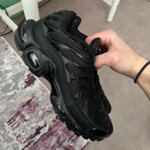 Nike Air Max Plus Triple Black - Nike Air Max Plus i helsvart färg med snygga vågformade detaljer och synliga Air-enheter i sulan. Ovandelen är i mesh och syntet, med svarta snören och diskret Swoosh på sidan. Riktigt clean och sportig look som sticker ut.