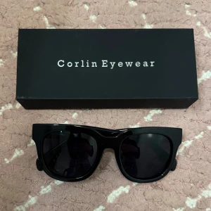Svarta solglasögon Corlin Eyewear - Snygga svarta solglasögon från Corlin Eyewear med bred båge och mörka glas. Modellen har en modern, kantig form som ger en cool vibe och passar perfekt till alla outfits. Kommer med originalask.