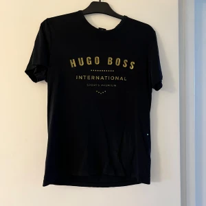 Svart Hugo Boss t-shirt med guldtryck - Svart t-shirt från Hugo Boss med guldig logga och text på bröstet. Klassisk rund halsringning och korta ärmar. Tillverkad i mjuk bomull som känns skön mot huden. Snygg och stilren design som lyfter din outfit.