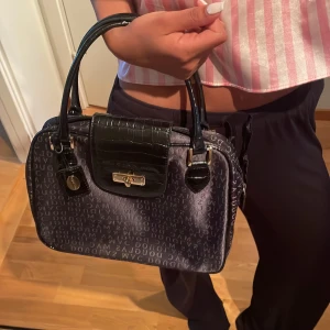 Svart handväska från Mac Douglas - Snygg svart handväska från Mac Douglas med glansig finish och präglat logomönster över hela väskan. Locket och detaljerna är i croco-mönstrat skinn med guldfärgat spänne. Två handtag och en liten tag med logga framtill.