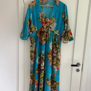 Blommig långklänning från Turquoise - Superfin långklänning i turkosblått från Turquoise med färgglatt blommönster. Klänningen har v-ringning, trekvartsärm och ett luftigt fall. Perfekt för dig som gillar boho-vibe och vill sticka ut med färg och mönster.