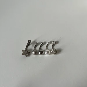 Navelpiercingar - Fem olika navelpiercings i silverfärg, en med blomformad kristall, en med glittrig kula, en med matt kula, en med pärlvit kula och en med glittrande stenar. 