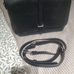 Svart axelväska med dragkedjor - Snygg svart axelväska med justerbar och avtagbar axelrem. Väskan har flera fack med dragkedjor både inuti och på baksidan, perfekt för att hålla ordning på dina grejer. Stilren design med silverfärgade detaljer och struktur i materialet.