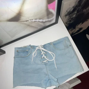 Ljusblå jeansshorts med snörning - Snygga ljusblå jeansshorts med hög midja och råa kanter. Framtill finns en cool snörningsdetalj med vita snören istället för dragkedja, samt klassiska fickor. Perfekta för sommaren och ger en trendig vibe.