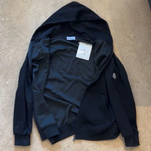 Svart hoodiejacka från Moncler L - Svart hoodiejacka från Moncler med dragkedja, huva med snörning och klassisk logga på ärmen. Jackan har känguruficka framtill och ribbade muddar. Perfekt för en chill och stilren look.