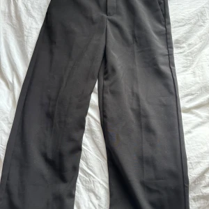 Svarta kostymbyxor med raka ben - Snygga svarta kostymbyxor med klassisk passform och raka, vida ben. Byxorna har hällor för bälte, diskreta fickor och en stilren look som funkar till allt. Perfekta för dig som gillar en clean och tidlös stil.