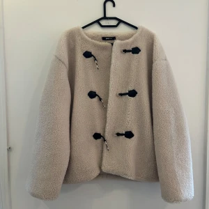 Beige teddyjacka från Gina Tricot - Supermjuk och fluffig beige teddyjacka från Gina Tricot med coola svarta detaljer och knäppning framtill. Jackan har rund hals, lång ärm och är fodrad på insidan för extra komfort. 