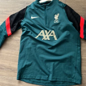 Liverpool Nike Dri-FIT träningsöverdel  - Liverpool FC träningsöverdel från Nike i Dri-FIT-material. Tröjan är mörkgrön med svarta partier på axlarna och röda detaljer på ärmarna. Halvlång dragkedja framtill, LFC-logga och AXA-sponsortryck på bröstet. Perfekt för fotbollsträning.