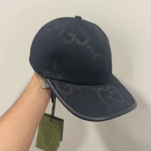 Svart keps från Gucci med GG-mönster - Snygg svart keps från Gucci med diskret GG-logga i ton-i-ton över hela kepsen. Klassisk böjd skärm och stilren design. Tillverkad i slitstark polyester med märkets gröna etikett kvar. Perfekt accessoar för att lyfta din outfit.
