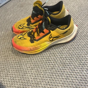 Nike löparskor Pegasus model - Nike löparskor i en riktigt fet gul och orange färg med svarta detaljer och snörning. Ovandelen är i mesh-liknande syntetmaterial för bra andning. Snygg svart swoosh på sidan och cool design på sulan. Perfekta för dig som vill sticka ut på löprundan.