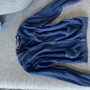 Marinblå kofta från GANT - Snygg marinblå kofta från GANT i storlek 13-14Y, passar dig som är 158-164 cm. Koftan har långa ärmar, knappar framtill och ribbade muddar. Perfekt att slänga över en t-shirt för en chill och klassisk look.