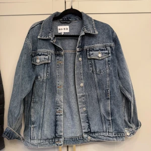 Oversized jeansjacka från NA-KD - Snygg oversized jeansjacka från NA-KD i klassisk blå denim. Jackan har långa ärmar, två bröstfickor med knappar och silverfärgade detaljer. Strl M