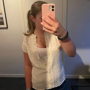 Vit stickad väst med knappar - Vit stickad väst med korta ärmar och snyggt hålmönster. Västen har stora knappar framtill, två små fickor och en v-ringad krage. Perfekt att styla över en t-shirt eller linne för en trendig look.