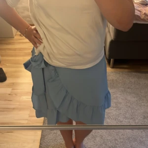 Ljusblå volangkjol från Vero Moda - Supersöt ljusblå kjol från Vero Moda med volangkant och omlottkänsla. Kjolen har en stor rosett i sidan och resår i midjan för extra bekvämlighet. Perfekt för dig som vill ha en feminin och trendig look i sommar.