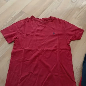 Röd t-shirt från Polo Ralph Lauren - Röd t-shirt från Polo Ralph Lauren med klassisk blå broderad logga på bröstet. T-shirten har rund halsringning och korta ärmar. Tillverkad i mjuk bomull som är skön att bära. Perfekt basic-plagg med stilren design.